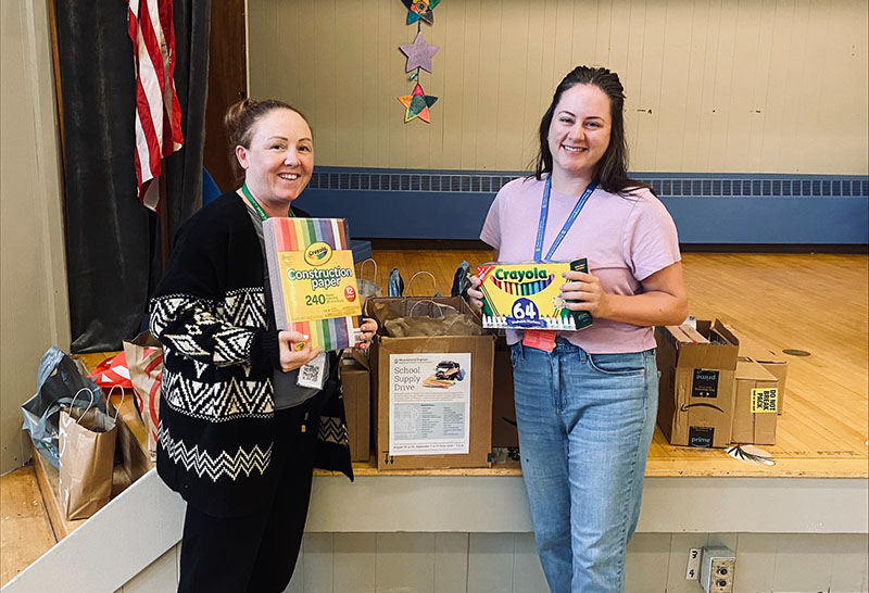 Two women standing in front of a collection of school supplies in a school gymnasium